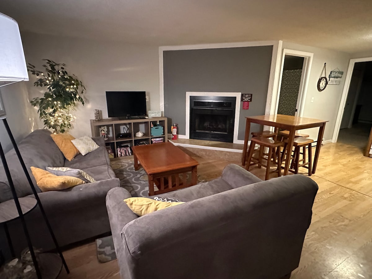 A cozy living room features a comfortable gray sofa and loveseat positioned around a wooden coffee table. A flat-screen TV is placed on a shelf stand next to a decorative plant. A fireplace with a dark surround is visible against the back wall, with a dining table and chairs in the background.