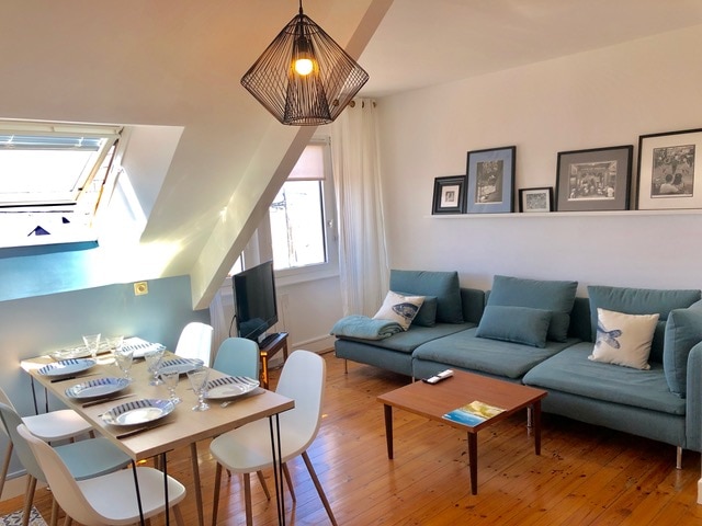 A bright living area features a modern sectional sofa paired with a wooden coffee table. A dining table is set for six, with plates and glasses arranged neatly. Natural light pours in from the skylights and windows, illuminating the light-colored walls and wooden flooring.