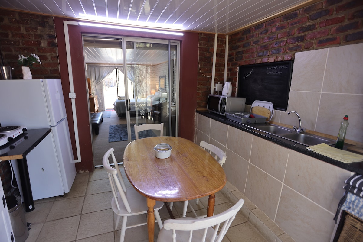 A well-equipped kitchen features a small table with four white chairs. The countertop holds a microwave, kettle, and several cooking essentials. A view of the garden is available through sliding glass doors, with a cozy sleeping area visible in the background.