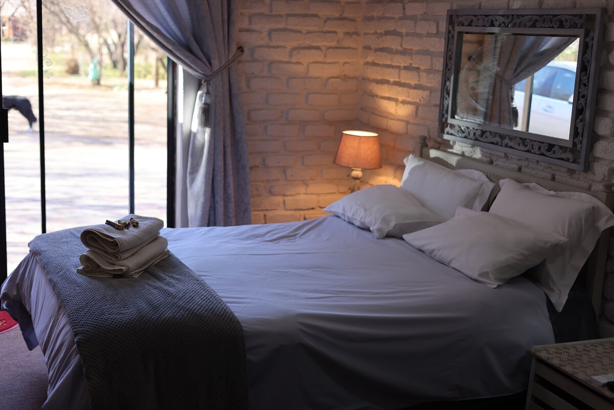 A bright and inviting bedroom features a queen bed with white linens. Soft pillows rest against the headboard, while a cozy blanket is draped over the bed. A bedside lamp illuminates the space, and large windows offer views of the outside.
