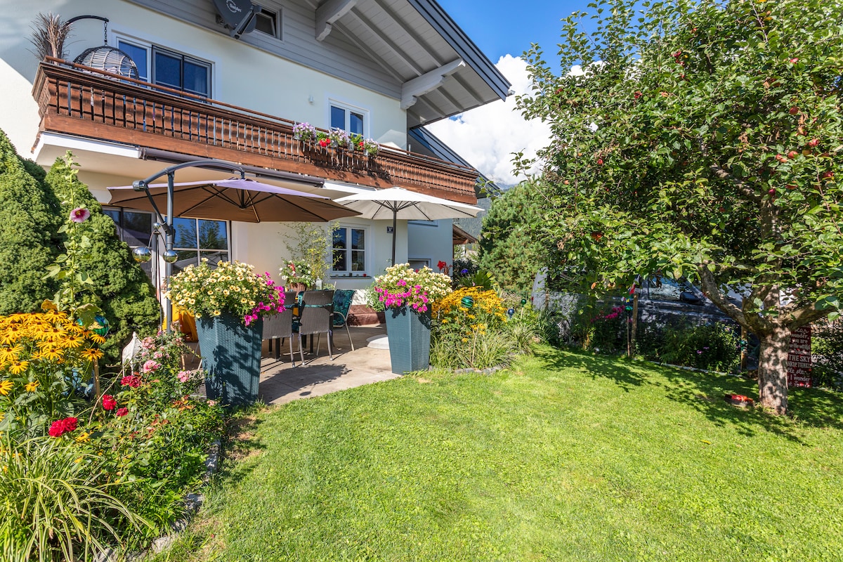 A well-maintained garden area surrounds a spacious patio. Sun umbrellas provide shade, with floral arrangements in large planters adding vibrant color. The residence features a balcony with floral boxes, enhancing the inviting outdoor space, which is lined with green grass and trees.