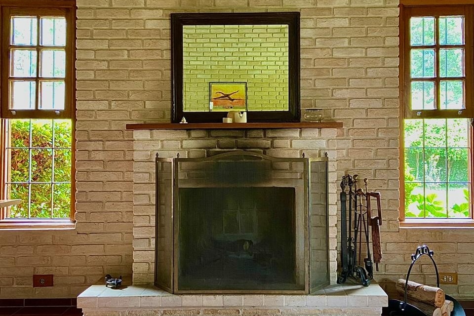 A warm interior features a brick fireplace framed by a wooden mantel. A large mirror hangs above the mantel, reflecting natural light from the nearby windows. The space is accentuated by greenery visible outside, creating a serene atmosphere.