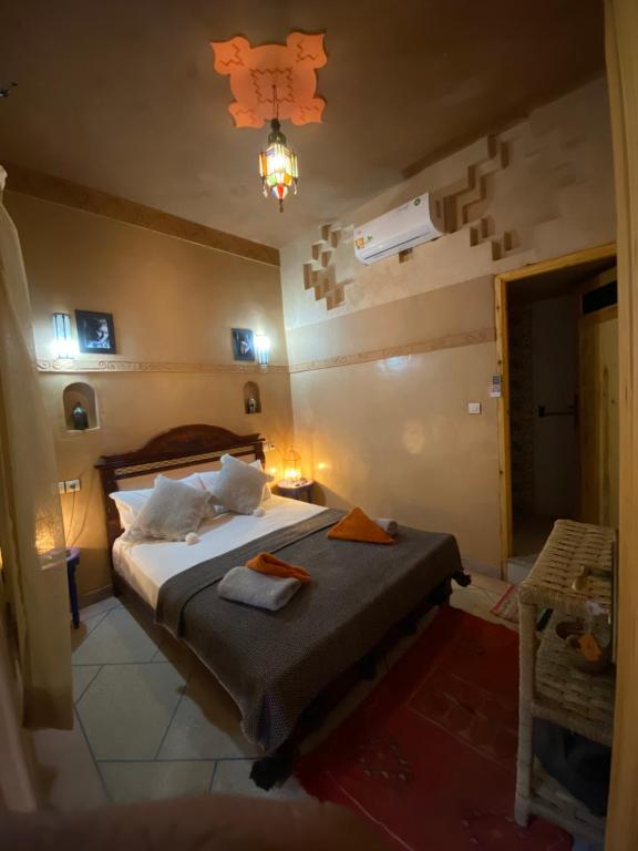 The room features a spacious double bed adorned with decorative pillows, complemented by a single bed nearby. Soft lighting creates a warm atmosphere, while traditional wall decor enhances the space. A small table and basket add functionality, and an air conditioning unit is visible.