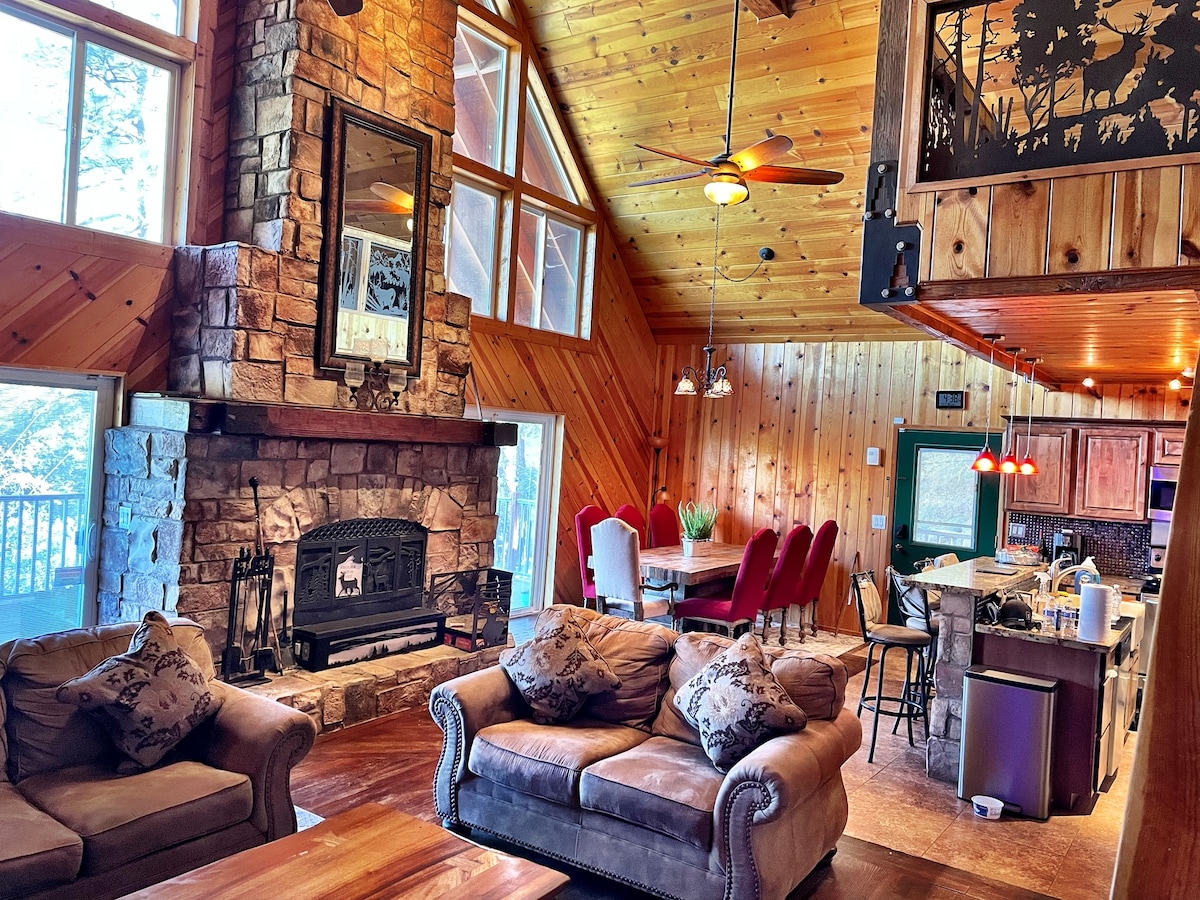 The living area features a stone fireplace framed by warm wooden walls and a high A-frame ceiling. Comfortable seating is arranged around a coffee table, while a dining area with a set of red chairs is positioned in the background, visible from the open kitchen space.