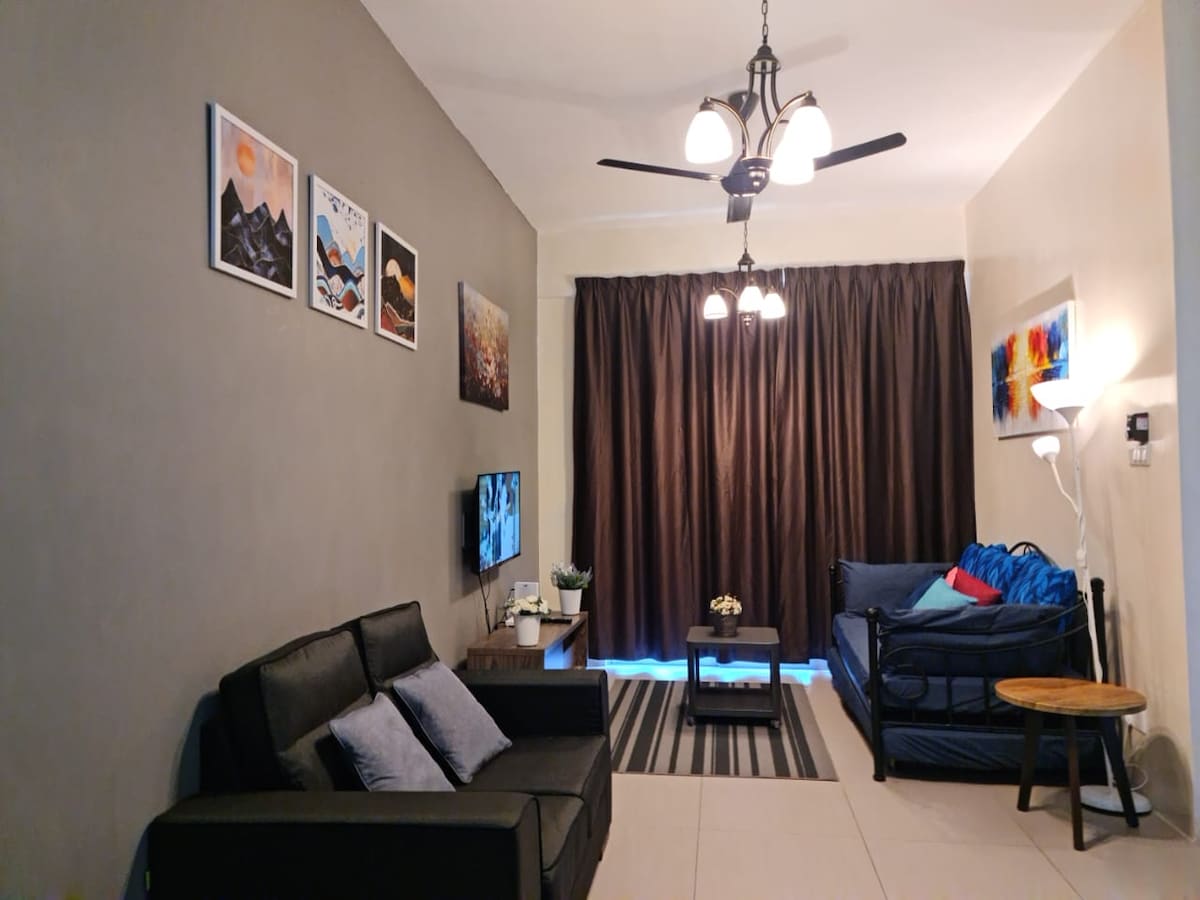 A living area is depicted with dark curtains covering large windows, allowing soft light to filter through. Two sofas, including a daybed, are positioned next to a small table. Wall art enhances the ambiance, while a television is mounted on the wall.
