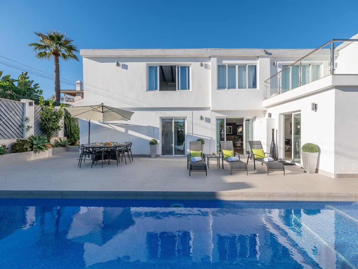The exterior of a modern villa is captured, featuring a shimmering pool with clear blue water. Sun loungers are arranged beside the pool, while a dining table is set under a shaded area. The villa showcases large glass doors and windows, surrounded by lush greenery.