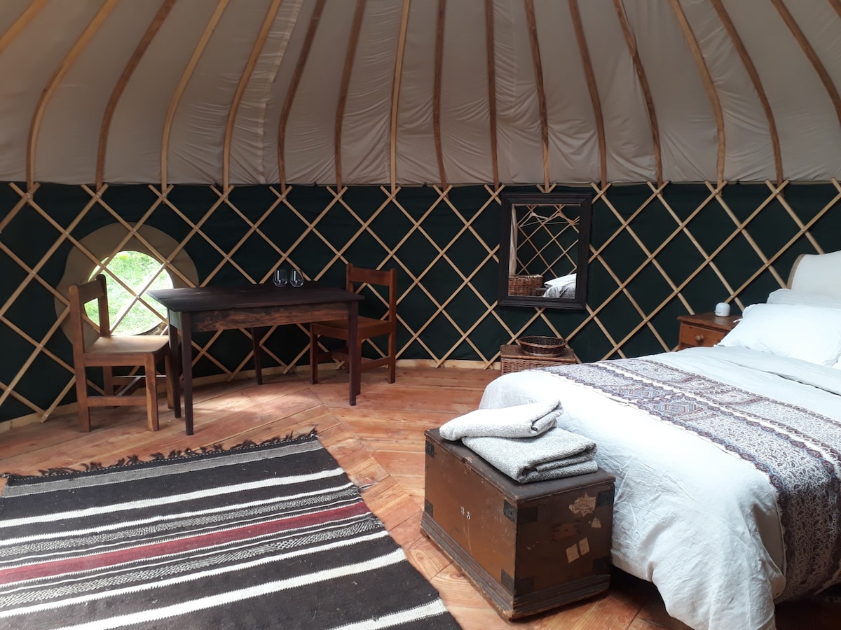 The interior of the yurt features a large double bed with crisp bedding and woolen throws. A simple wooden table and chairs are positioned nearby, alongside a rustic chest. A round window offers natural light, while a small mirror is mounted on the wall.