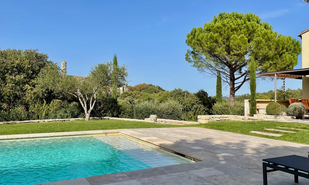 A clear view of the saltwater pool reflects the sky, framed by smooth stone decking. Lush greenery, including a prominent tree, surrounds the area, enhancing the serene ambiance. In the background, hints of the historic architecture add contrast to the natural landscape.