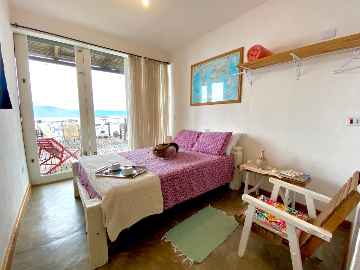 A cozy sleeping area features a spacious bed covered with a patterned blanket. Natural light floods in from glass doors that lead to a private deck, furnished with vibrant outdoor chairs. A small table and decorative items complete the inviting interior.