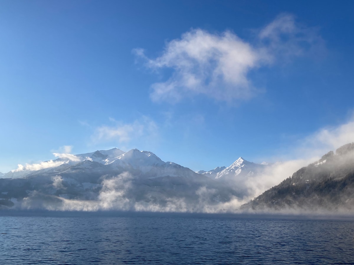 A scenic view features snow-capped mountains against a clear blue sky. Wisps of fog hover over the lake's surface, creating a serene atmosphere. The landscape includes hints of greenery along the lake's edges, complementing the tranquil waters below.