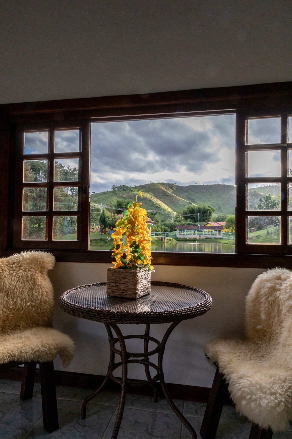 A cozy seating area features a round wicker table surrounded by two chairs with soft coverings. A bright arrangement of yellow flowers sits in a basket at the center. The window offers a view of a mountainous landscape and a tranquil lake.