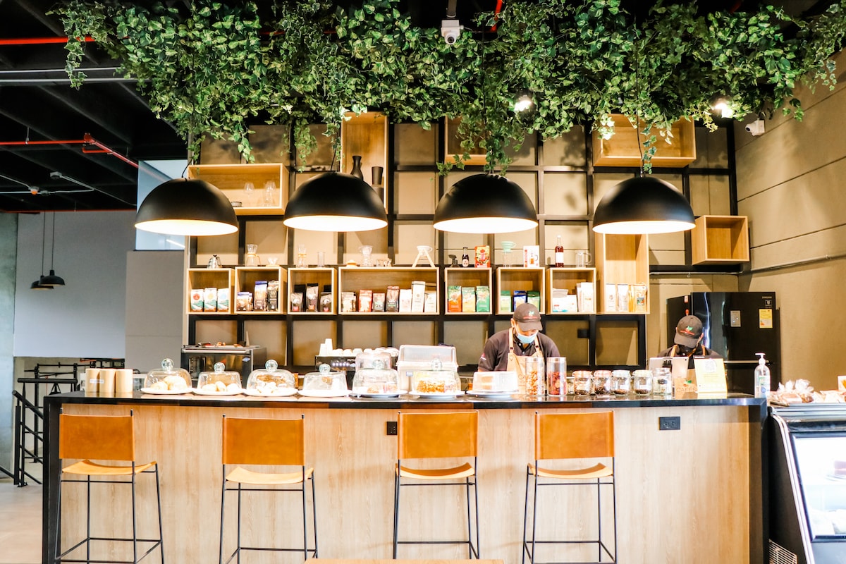 A modern snack bar is showcased, featuring a curved wooden counter with bar stools. Dark pendant lights hang from the ceiling, and a wall of shelves displays various snacks and beverages. Green foliage decorates the upper area, creating a refreshing atmosphere.