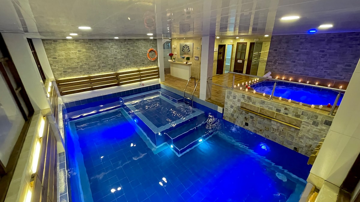 An indoor pool area features a main swimming pool with stairs leading into the water, alongside a smaller hot tub. The space is illuminated with soft lighting, and the walls are adorned with stone accents, creating a serene atmosphere. Wood and stone elements offer a contemporary touch.