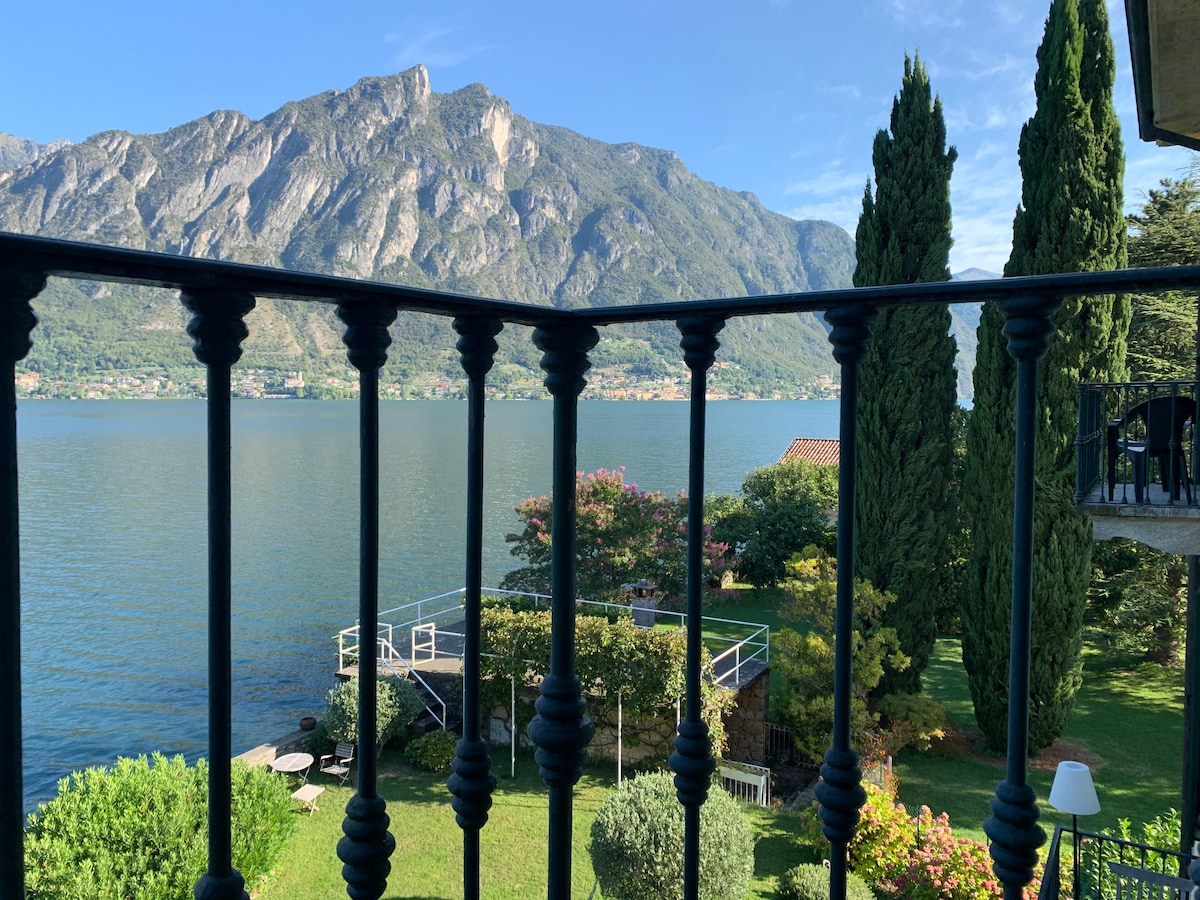The image displays a view from a small balcony overlooking a serene lake, framed by tall cypress trees. Mountains rise majestically in the background, while a well-maintained garden with a variety of greenery is visible below, enhancing the natural setting.