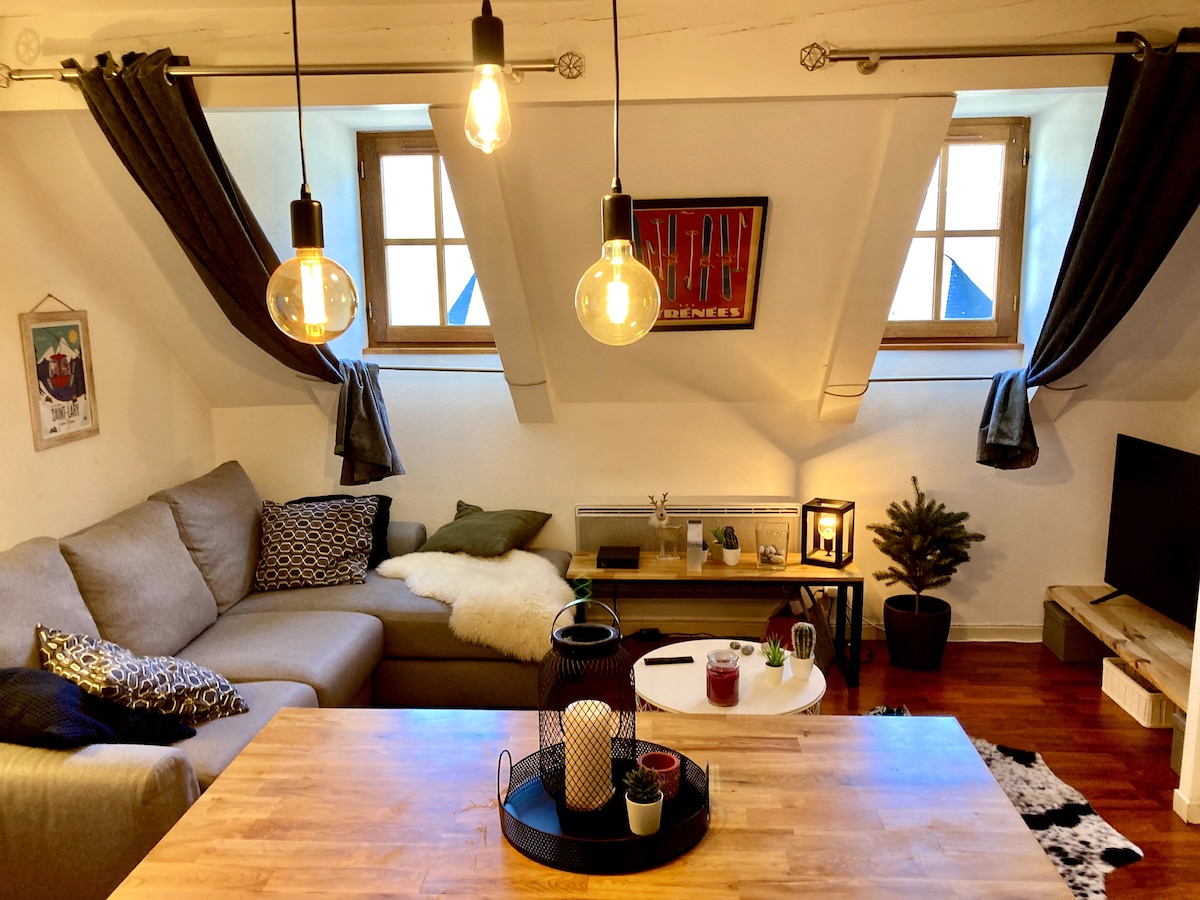 A cozy living area features a neutral sofa with decorative cushions, complemented by a small coffee table. Ambient lighting is provided by hanging bulbs, while two windows allow natural light to illuminate the space. A decorative plant and artwork add subtle character.