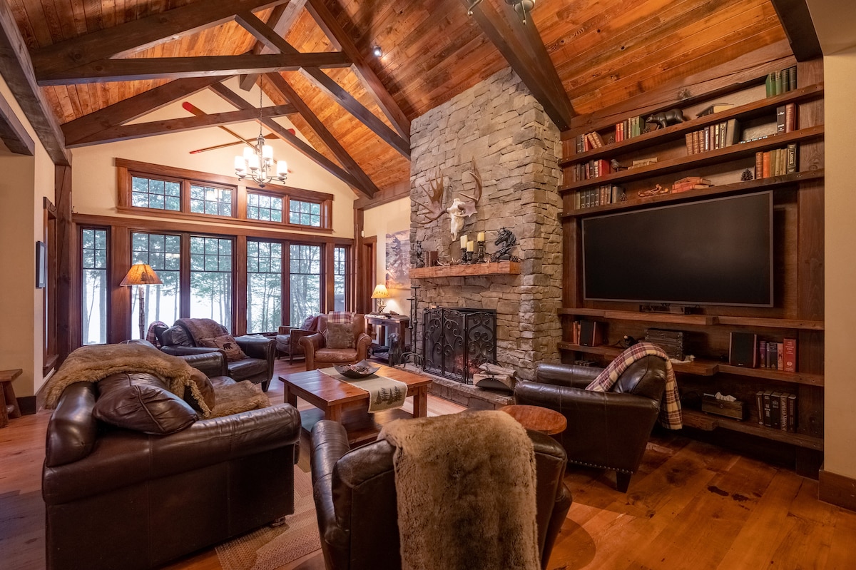 A spacious living area features a high, wood-beamed ceiling and a stone fireplace as a focal point. Comfortable leather sofas surround a central wooden coffee table, while large windows allow natural light to filter in, offering views of the outdoor landscape.