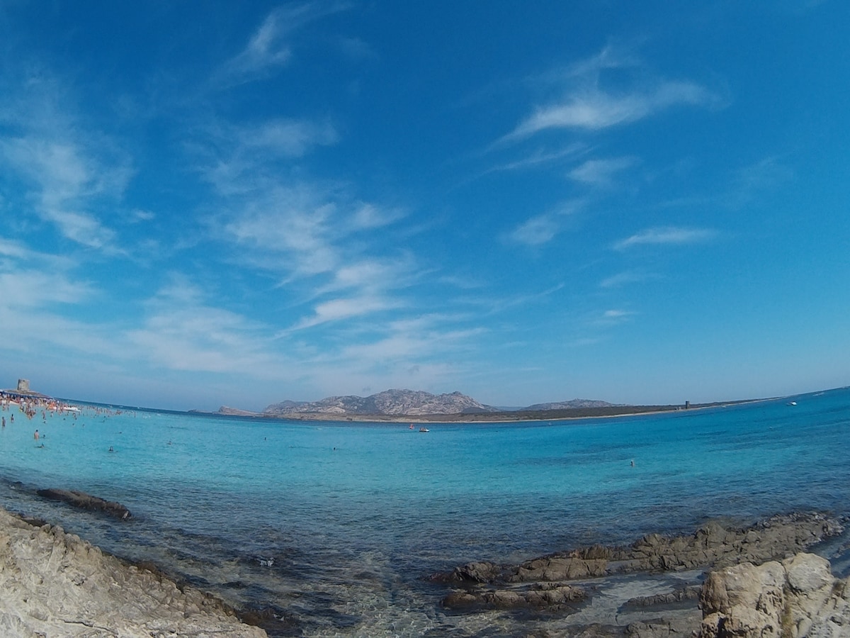 The image presents a panoramic view of a serene beach with crystal-clear turquoise waters. Rocky shores create a natural boundary, while distant islands are visible on the horizon under a vibrant blue sky with scattered clouds. Sunbathers can be seen enjoying the shoreline.