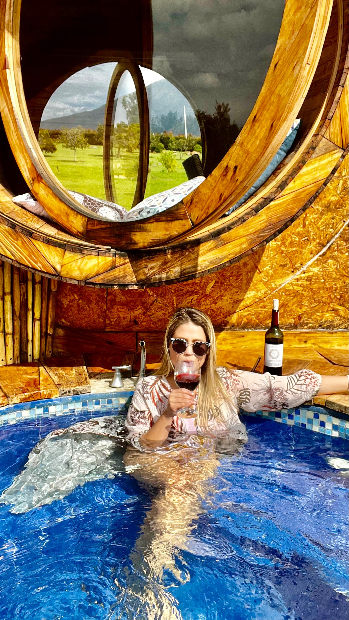 A circular window frames a scenic view of the outdoors, with mountains visible in the distance. A cozy, private jacuzzi is filled with water, reflecting natural light, while a bottle of wine is placed nearby. Inviting colors and textures enhance the tranquil setting.