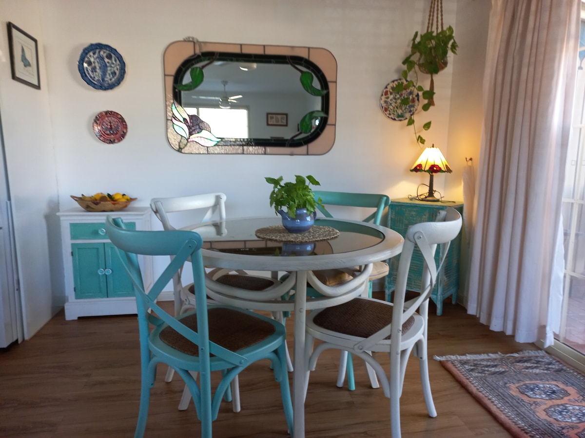 A dining area is portrayed, featuring a round table surrounded by four chairs. A small potted plant sits at the center of the table, while colorful plates adorn the walls. A mirror reflects the light from a nearby lamp, enhancing the inviting feel of the space.