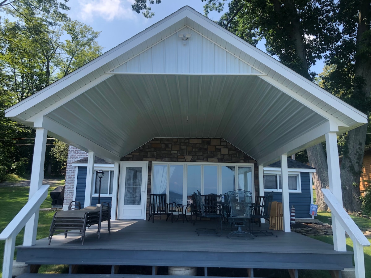 A covered outdoor deck features a combination of seating options, including several chairs and a glass-top table. The back of the lakefront home is visible through large glass doors, enhancing the connection between indoor and outdoor spaces. Lush trees frame the area, providing shade.