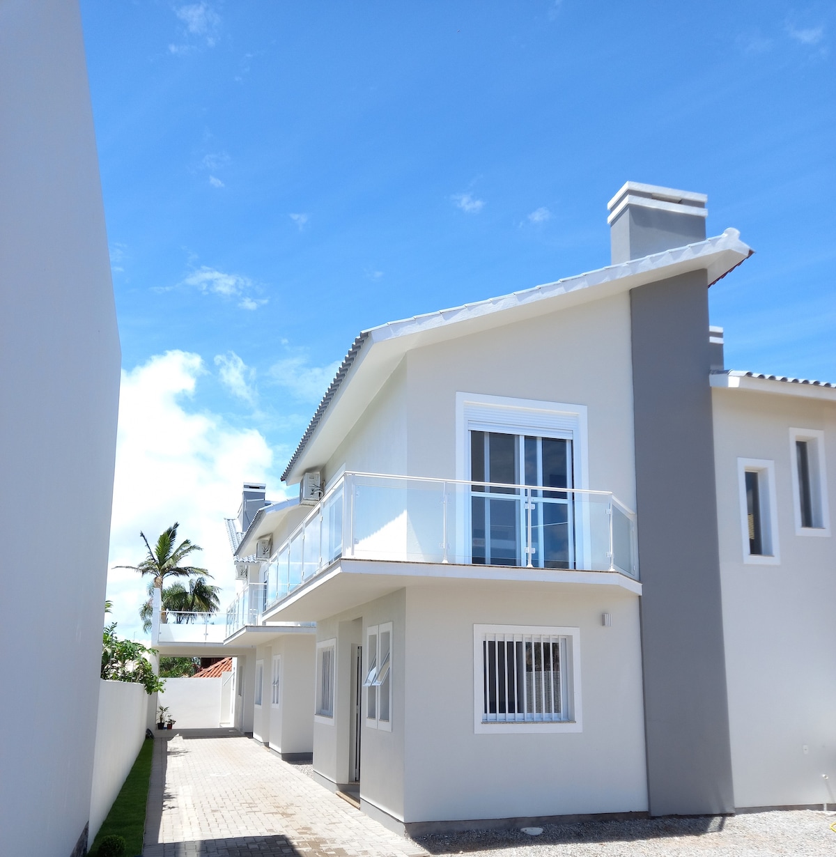 The exterior of a two-story house is depicted, featuring a light-colored façade and a balcony on the second level. A pathway leads to the entrance, flanked by greenery, while palm trees are visible in the background under a clear blue sky.