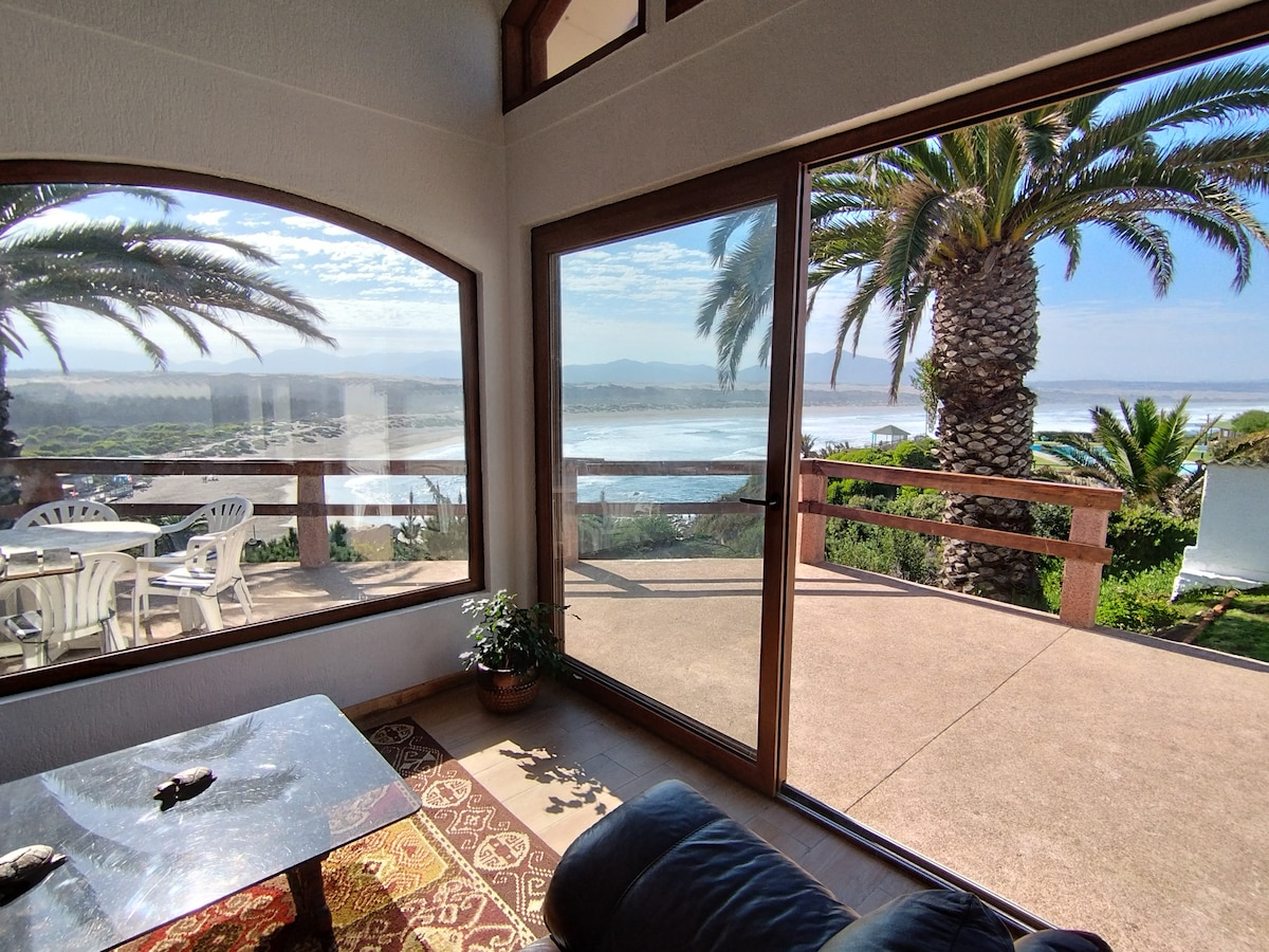 A spacious living area is highlighted by large arched windows, offering a panoramic view of the coastline and palm trees. A dark sofa is positioned near the glass doors, with a small table visible in front. Natural light fills the space, creating a bright environment.