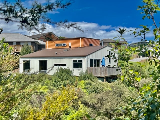 The modern split-level home is set among lush greenery, featuring a combination of light-colored and warm wooden siding. Large windows provide a glimpse of the interior, while a deck extends outward, complete with an umbrella. The surrounding landscape showcases native plants and shrubbery.