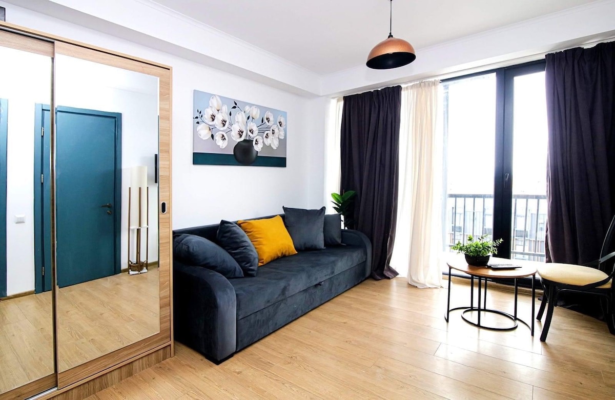 A modern living area features a dark sofa adorned with yellow cushions, positioned beside a small round table and two chairs. Large windows invite natural light, while sheer and opaque curtains provide varying degrees of privacy. A mirror reflects the room's design elements.