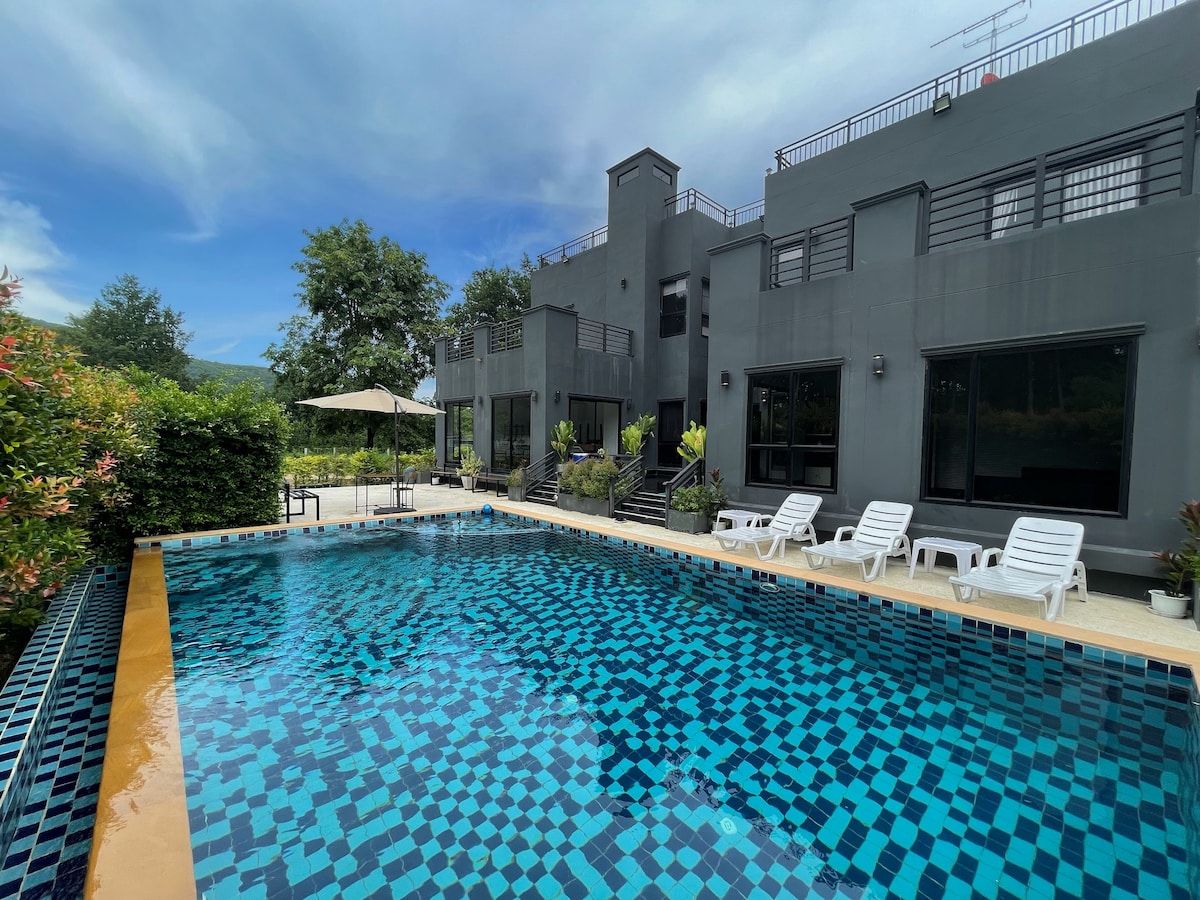 A modern villa features a spacious outdoor area with a 12-meter swimming pool, surrounded by colorful tiles. Several sun loungers are positioned around the pool, with a shaded seating area nearby. Lush greenery and mountains can be seen in the background, under a partly cloudy sky.