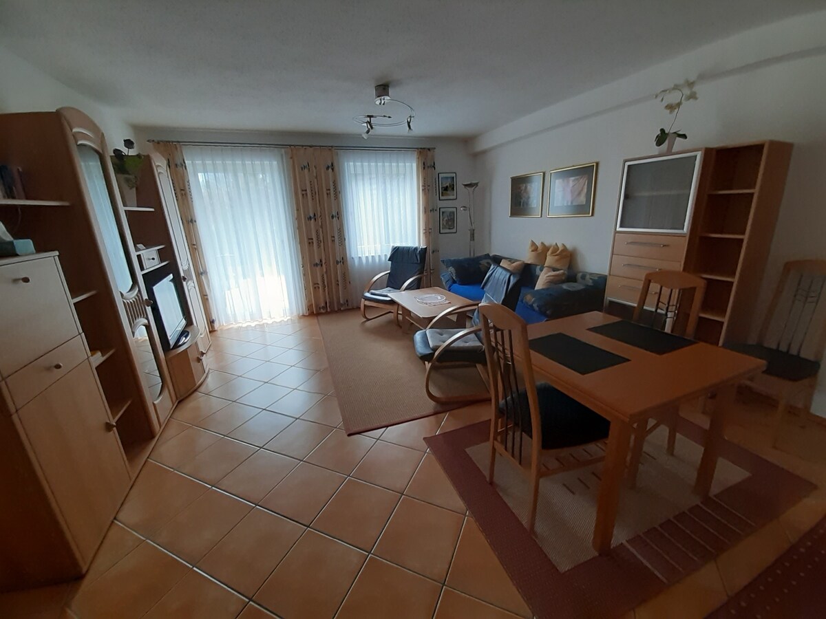 A bright living area features a comfortable seating arrangement with a sofa and chairs, alongside a wooden dining table with chairs. Large windows adorned with sheer curtains allow natural light to fill the space, enhancing the warm ambiance of the room with tiled flooring.