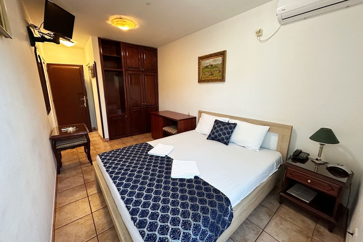 A well-lit room features a spacious bed adorned with white linens and a patterned navy blue throw. A desk and chair are positioned nearby, complemented by wooden cabinets. Additional details include a small bedside lamp and a flat-screen TV mounted on the wall.