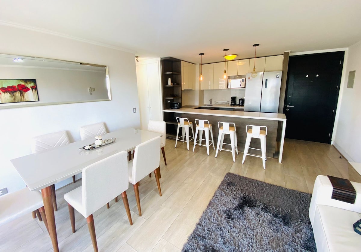 A modern dining area features a sleek white table paired with four light-colored chairs. Across from it, a contemporary kitchen showcases a spacious countertop and bar seating for four. Pendant lighting adds warmth to the room, complemented by a soft area rug underfoot.