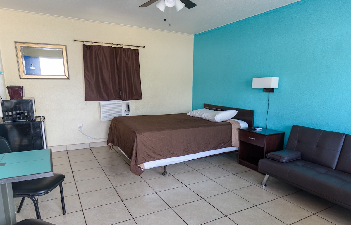 A spacious room features a neutral-colored bedspread covering a large bed, accompanied by standard pillows. A futon is positioned to the side, and a table with chairs is visible. A window air conditioning unit and a glass door mirror can be found, providing functionality.