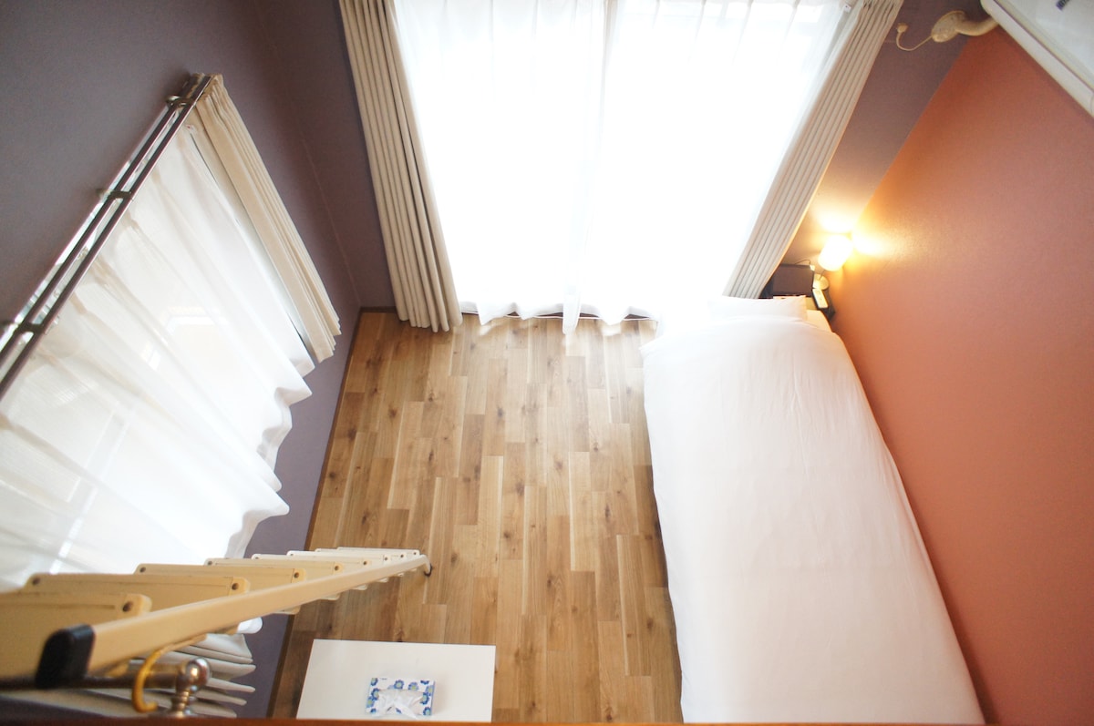 An overhead view showcases a cozy sleeping area with a spacious bed against a warm orange wall. Light enters through sheer curtains, illuminating the wooden floor. Additional sunlight filters in, creating a bright and airy feel in the room.