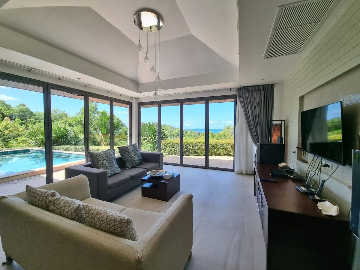 A modern living area features large glass doors that overlook a private pool and the lush greenery beyond. Comfortable seating is arranged around a low table, with light-colored sofas contrasting against the sleek furnishings. Natural light fills the space, creating an open and airy atmosphere.