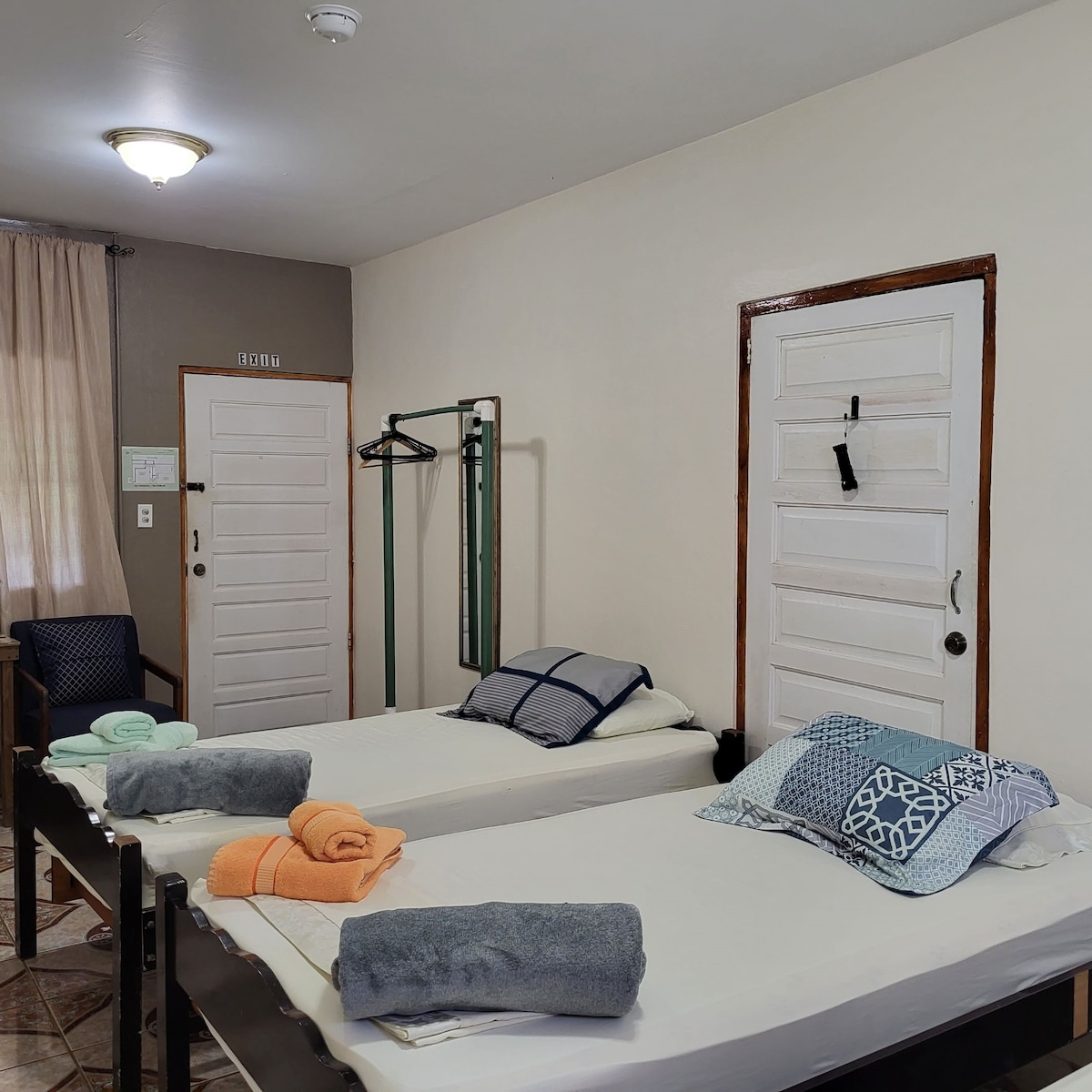 The image displays a spacious bedroom with two neatly made beds, each topped with colorful towels. A wooden chair is positioned beside a window, adorned with light curtains. A coat rack stands beside a closed door, while another door leads outside.