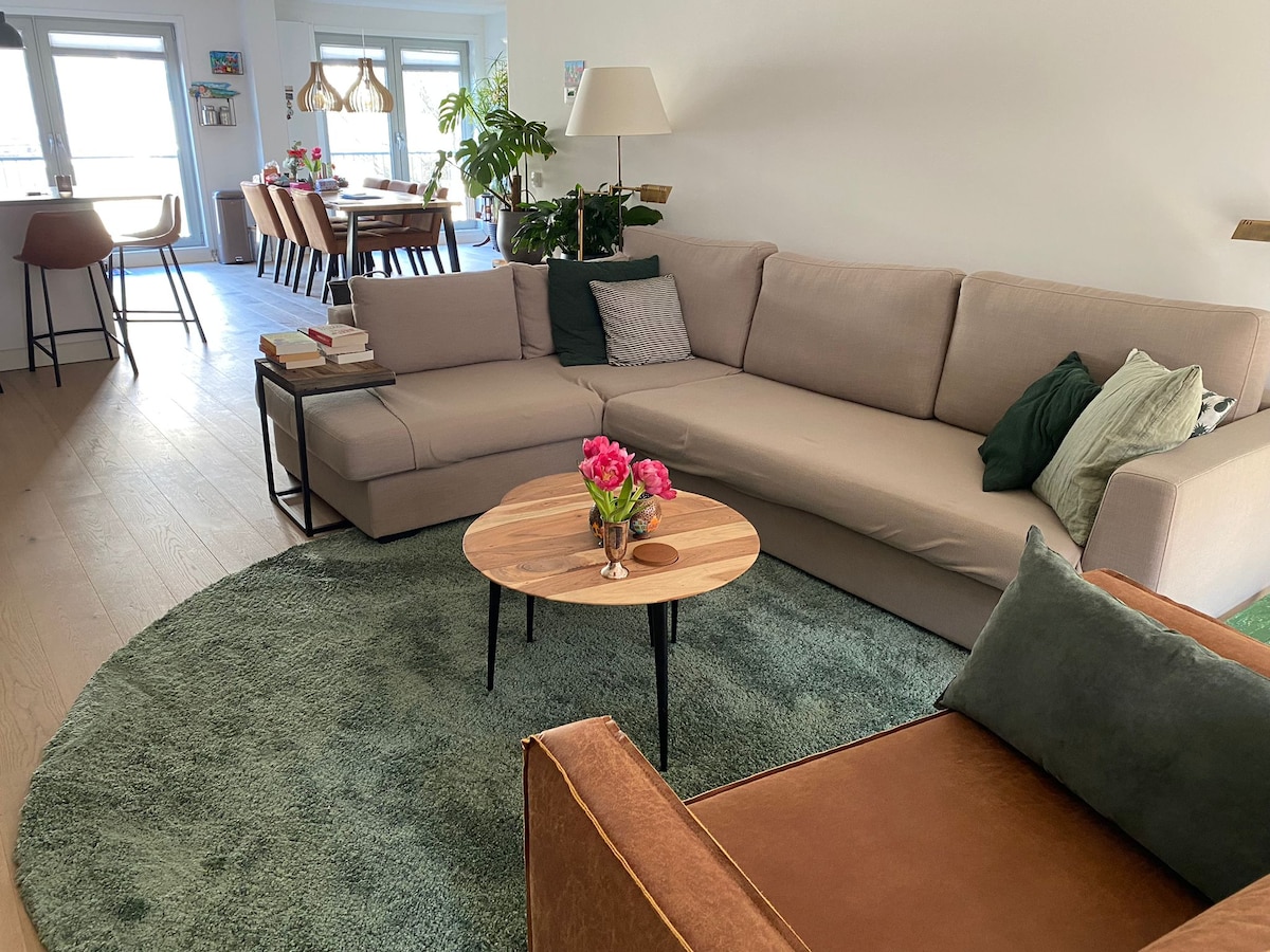 A spacious living area features a light-toned sectional sofa alongside a round coffee table and a green area rug. Natural light enters through large windows, while a dining table is visible in the background. Decorative plants add a touch of warmth to the space.