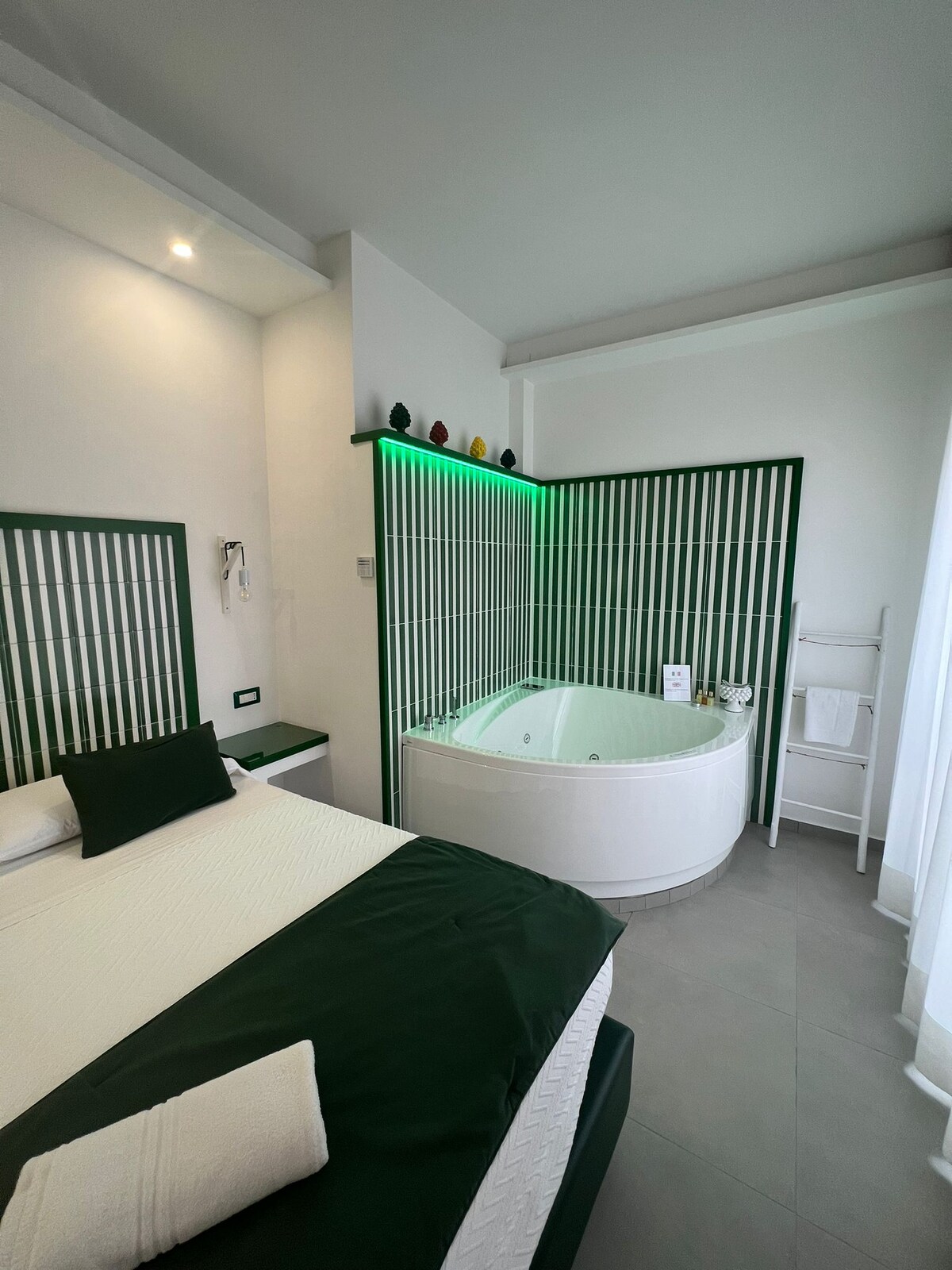 A cozy bedroom is highlighted by a spacious bed with green accents. Adjacent to the bed, a modern whirlpool tub is set within a sleek partition. Subtle lighting enhances the clean lines of the room, which is designed with neutral tones and minimalistic decor.