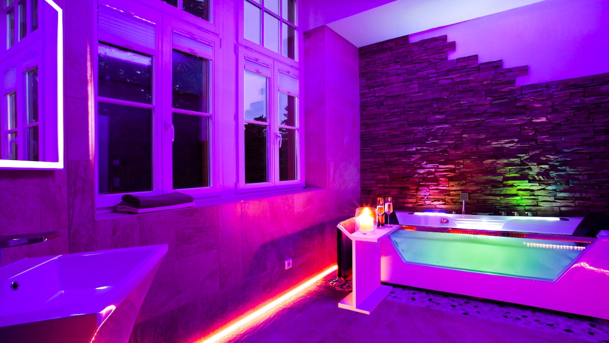 A spacious bathroom features a modern whirlpool tub, illuminated with soft purple and green lighting. Natural stone accents enhance the decor, while large windows allow natural light. A separate freestanding bathtub sits nearby, adding to the luxurious ambiance.