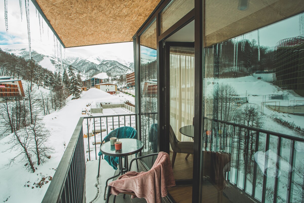 A balcony with a metal railing offers a small round table and two chairs, surrounded by snow-covered terrain. Icicles hang from the roof above. Majestic mountain views are reflected in the glass door, creating a serene winter ambiance.