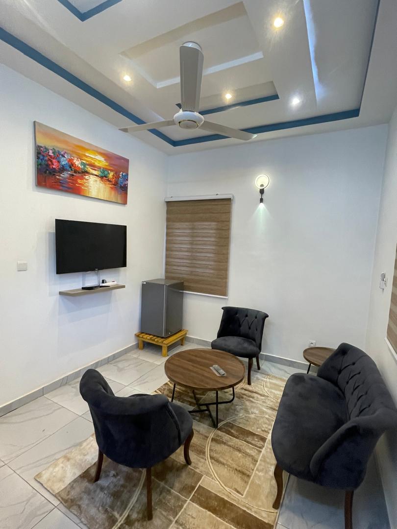 The living area features a flat-screen television mounted on the wall, with a small refrigerator visible nearby. Two comfortable chairs and a circular coffee table are positioned on a textured rug, while natural light enters through the window covered by beige blinds.