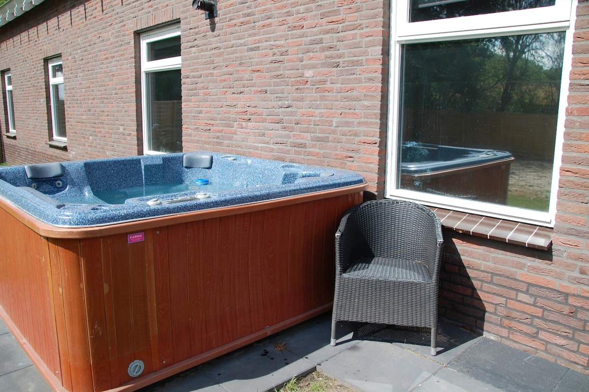 A private jacuzzi is positioned against a brick wall with a window reflecting its image. A wicker chair complements the relaxing space. The stone floor enhances the outdoor seating area, suitable for enjoying the tranquil surroundings.