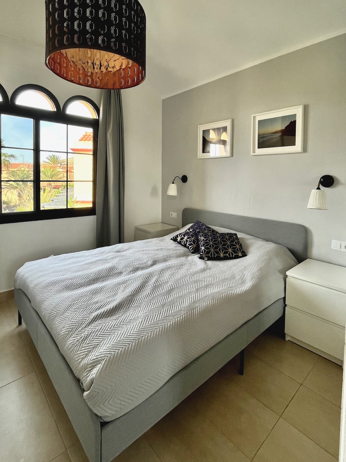 A bright bedroom features a spacious bed dressed in light bedding and decorative pillows. Two bedside tables are positioned on either side of the bed. A large window with arched design allows natural light to fill the space, complemented by two wall-mounted lamps and framed wall art.