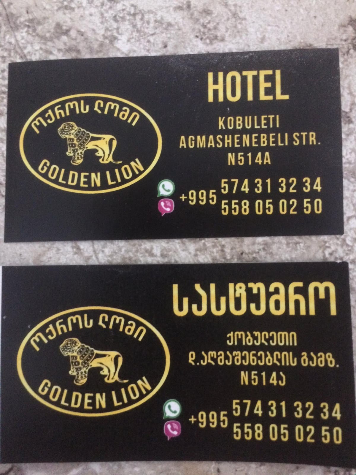 Two business cards are displayed, featuring the name and logo of the Golden Lion Hotel. The top card displays contact information and the hotel's location, while the bottom card provides additional details in Georgian. The black background contrasts with the gold text, creating a clear and professional appearance.