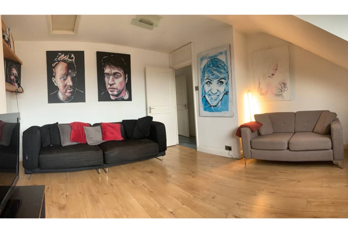 A bright lounge area features two stylish sofas in contrasting colors, adorned with colorful cushions. Light wood flooring enhances the space, which is complemented by wall art and a soft glow from a nearby lamp. A door leads to additional rooms.