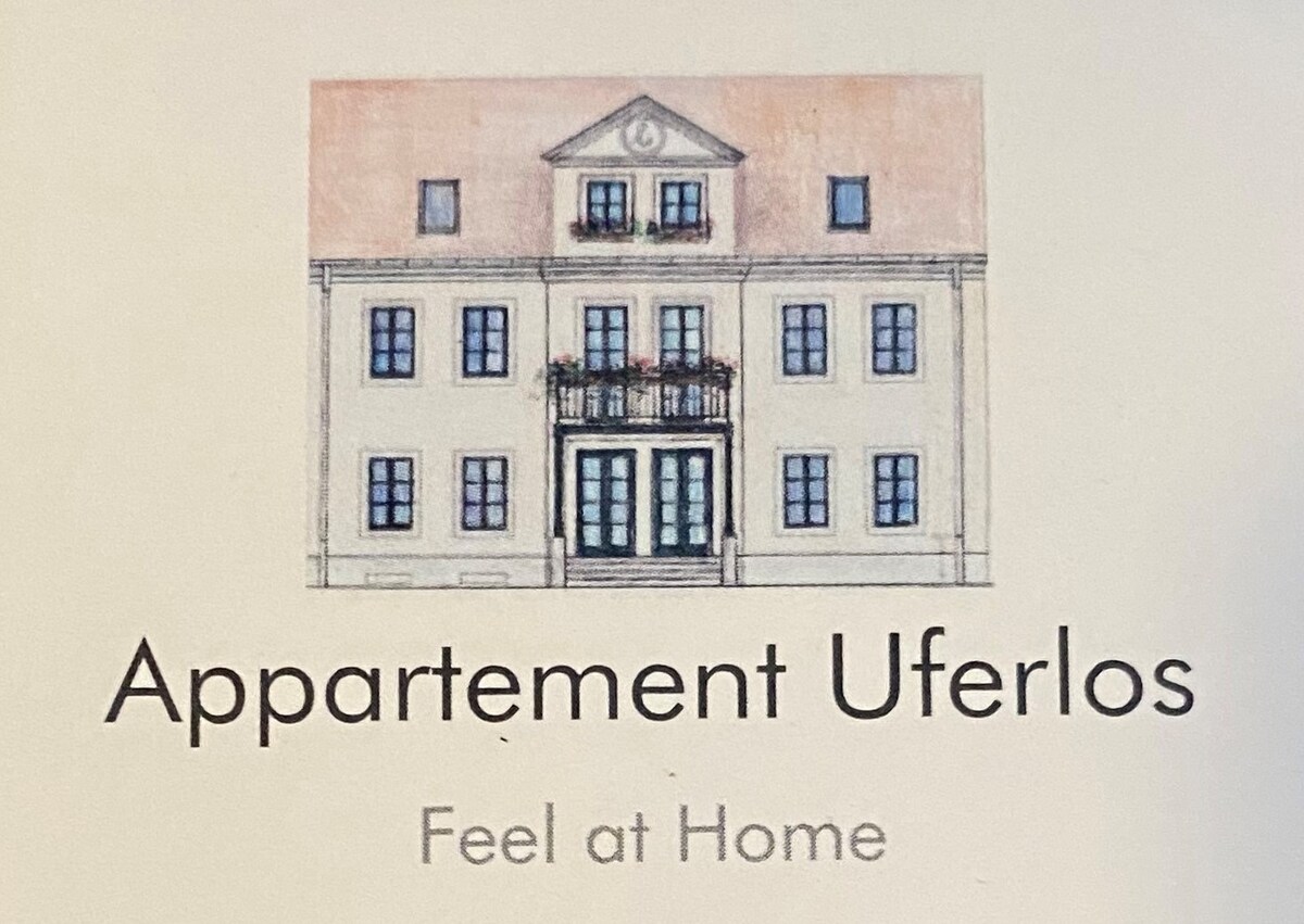 The facade of Appartement Uferlos is presented, highlighting a symmetrical two-story building with a central entrance framed by double doors. The structure features a balcony adorned with decorative flower boxes, with large windows that invite natural light into the interior.