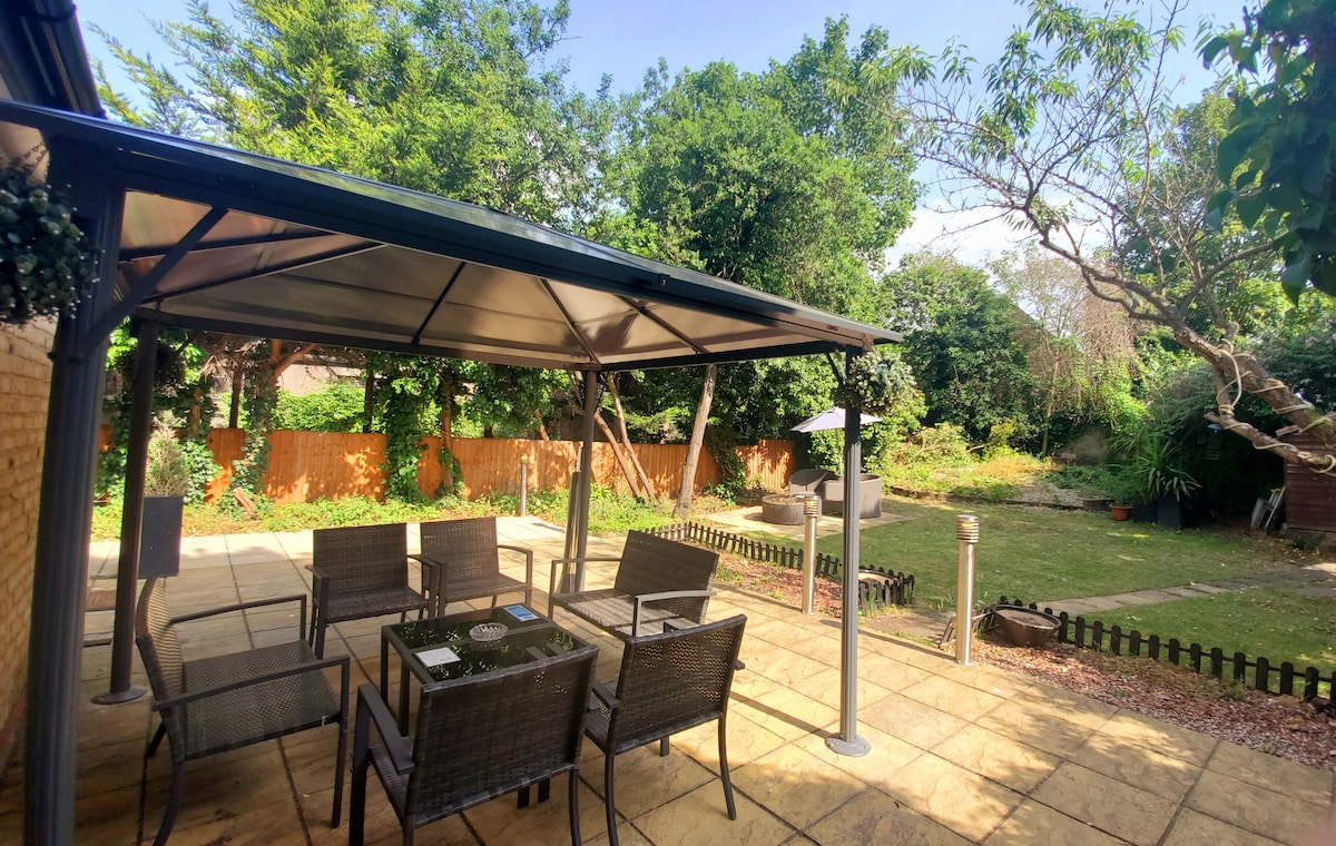 An outdoor seating area is shaded under a sturdy canopy, featuring a large table surrounded by several chairs. Lush greenery and trees create a natural backdrop, while a well-manicured garden area is visible in the distance, inviting relaxation and outdoor dining.