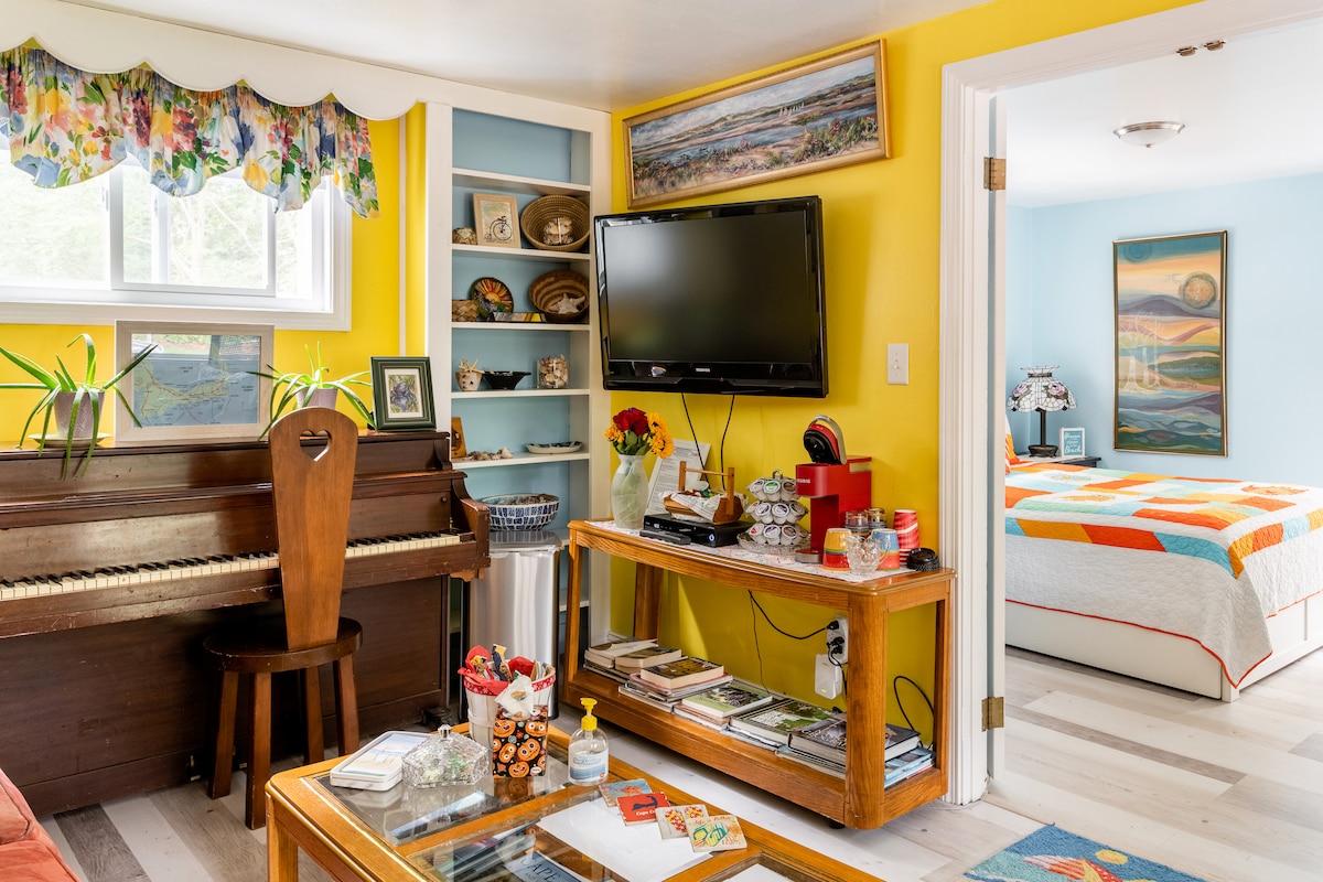 A cozy living area is featured, displaying a wooden piano and a television. Bright yellow walls are adorned with floral curtains. Shelves with decorative items and books are visible. A colorful quilt covers the bed in the adjacent room, enhancing the inviting environment.