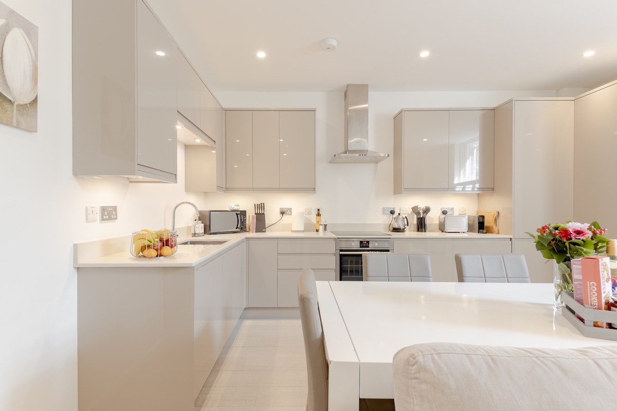 A modern kitchen is presented with sleek grey cabinetry and a white countertop. Appliances include a built-in oven, stovetop, and microwave. A dining table is visible with six chairs, and a fruit bowl is placed on the counter, accompanied by a bouquet of flowers on the table.
