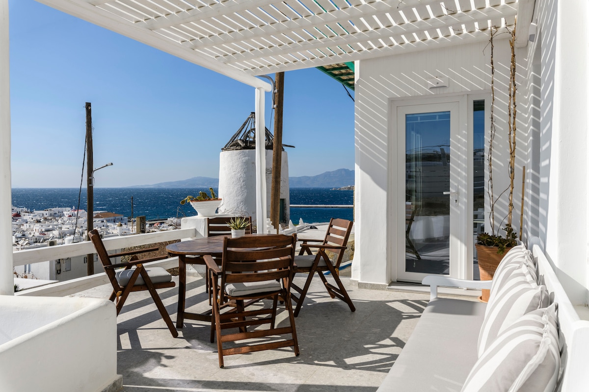 An outdoor terrace features elegant wooden furniture, including a round table surrounded by four slatted chairs. A cozy bench with light-colored cushions sits against the wall, providing a relaxing spot. The backdrop showcases stunning views of the Aegean Sea and Mykonos town.